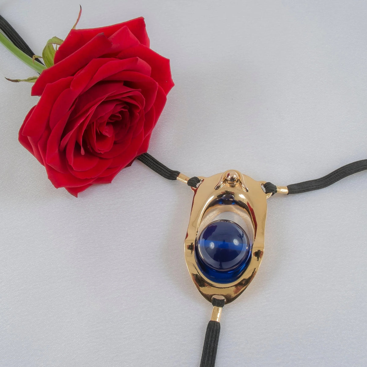 Goldener Klitorisschmuck mit blauem Cabochon auf schwarzer Kordel, elegant präsentiert neben roter Rose